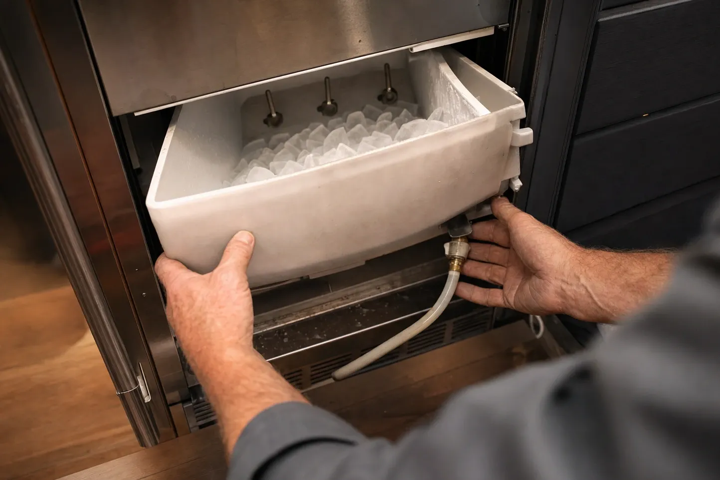 Hands lifting a white bin of cubes out of an undercounter ice maker, three fill spouts above the bin and a hose running from a brass fitting below it