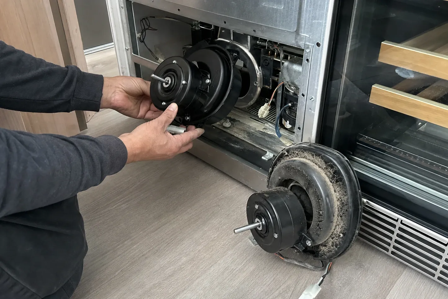 Hands holding a clean replacement fan motor at the open lower bay of a glass-door wine cabinet, the removed motor thick with dust standing on the floor
