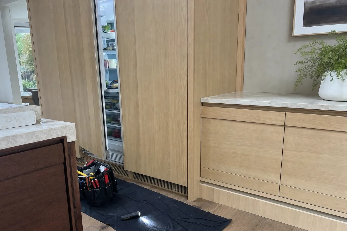 An oak-panelled integrated refrigerator column standing ajar in a run of flush cabinetry, stocked shelves lit inside, a tool bag and a lit torch on a drop cloth